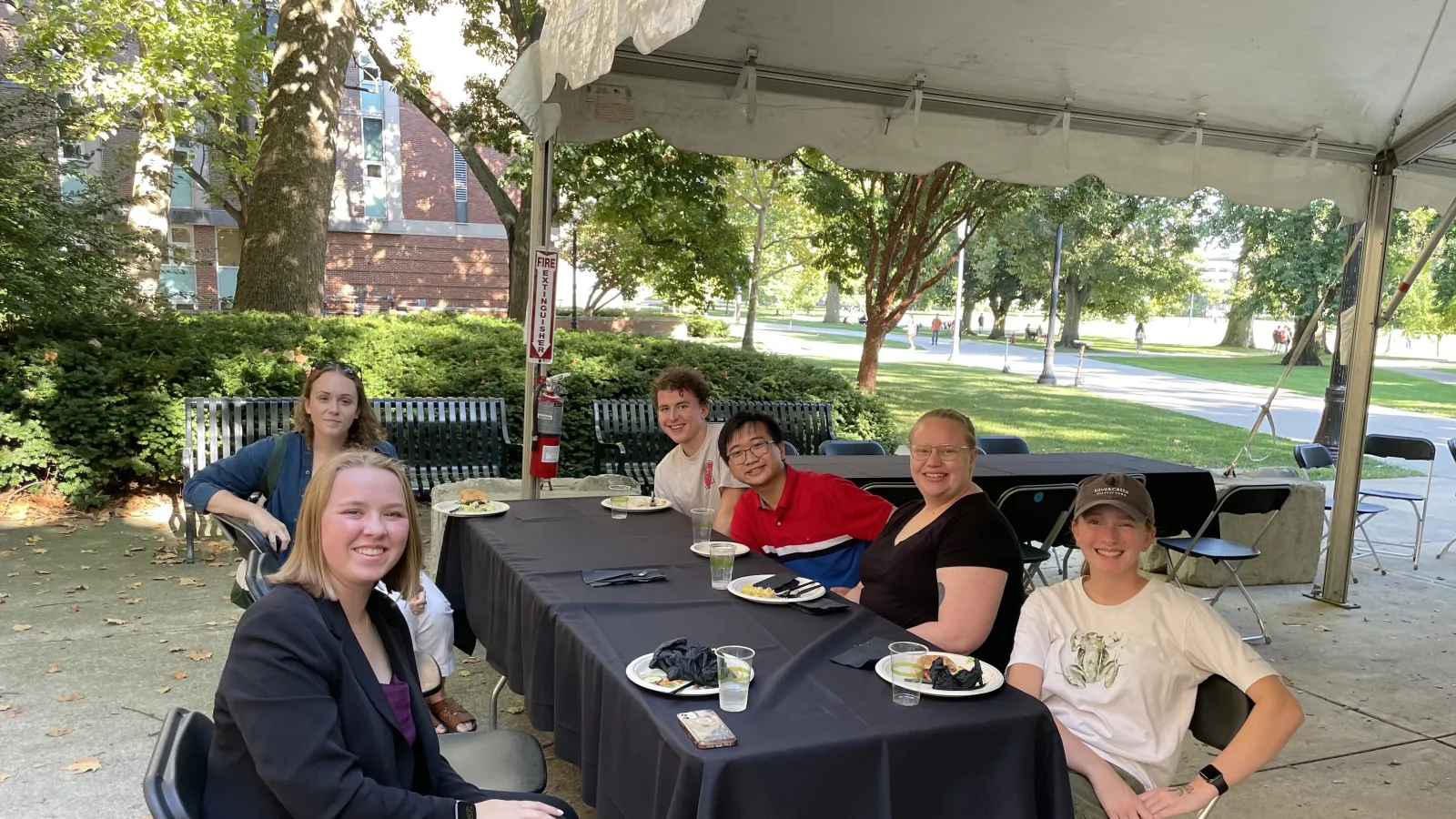 Group of 6 students sitting under at a table under a tent outside