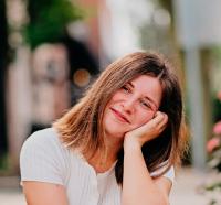 girl with hand on her face as she sits on the ground in white shirt in front of camera smiling