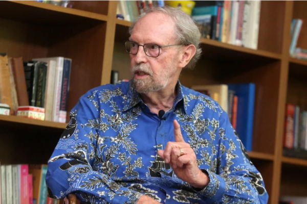 William Liddle, a white older man in a blue shirt talking in front of a bookcase