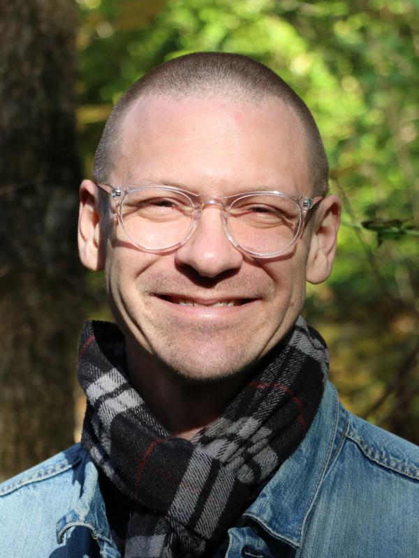William Minozzi headshot- white man in front of a tree wearing a denim shirt and plaid scarf